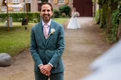Man in a green suit with a pink tie standing outdoors, with a woman in a white dress in the background.