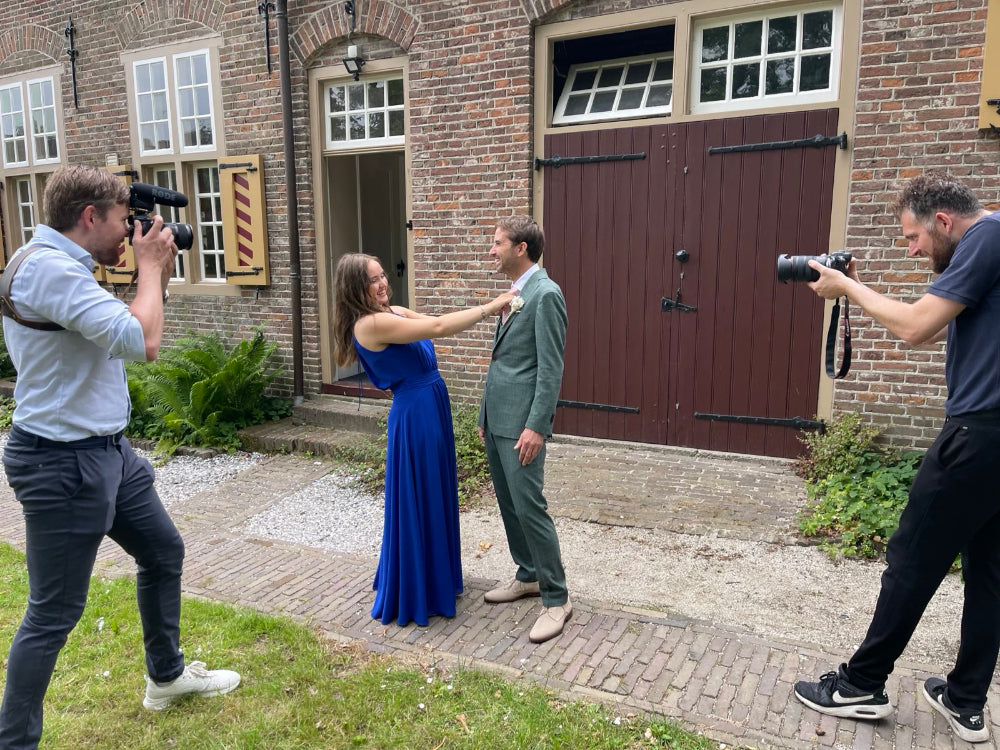 Two people in formal attire being photographed by two photographers in front of a brick building.