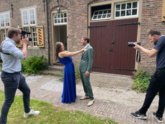 Two people in formal attire being photographed by two photographers in front of a brick building.