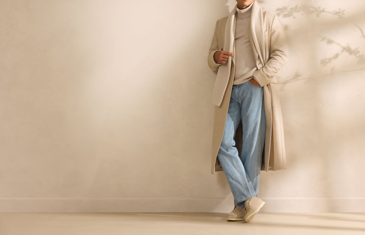 Model in a cream overcoat, ribbed scarf and light denim, leaning against a wall